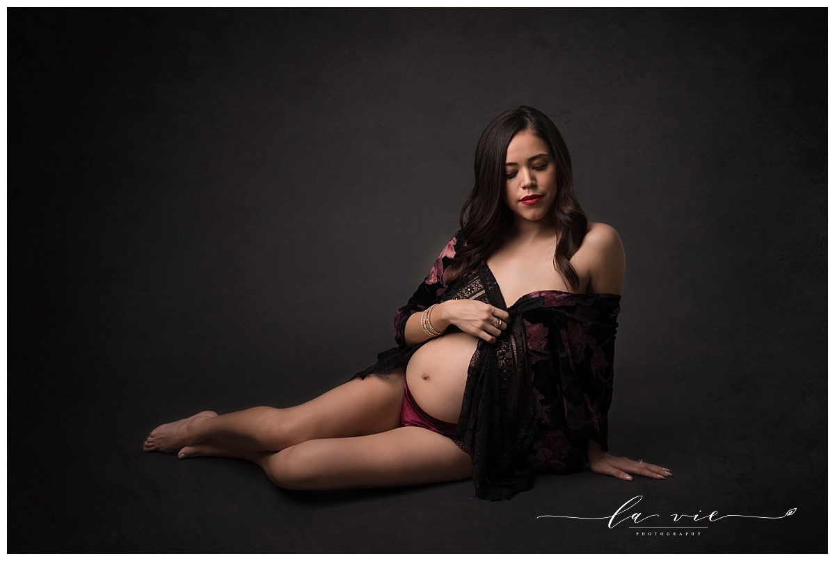 Reclined maternity pose with dark background wearing purple, red, and black lingerie
